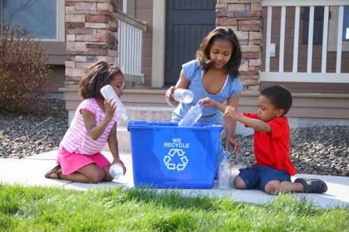 A young woman with shoulder-length curly hair, wearing a blue top, is standing on a sidewalk in front of a residential house with brick and stone exterior walls, a dark grey door, and a white railing. She is emptying plastic bottles into a large blue recycling bin marked with a white recycling symbol and the words 'WE RECYCLE.' Two children, a girl in a pink and white striped top and matching pink shorts, and a boy in a red top and dark shorts, are sitting on the grass nearby, holding and preparing more plastic bottles to add to the bin. The scene takes place on a bright, sunny day, with a well-maintained lawn, a concrete pathway, and a rock mulch bed with small bushes and plants, reflecting an environment focused on eco-friendly waste separation and rubbish management at a suburban residence, consistent with services provided by House Clearance Mottingham in the local area.
